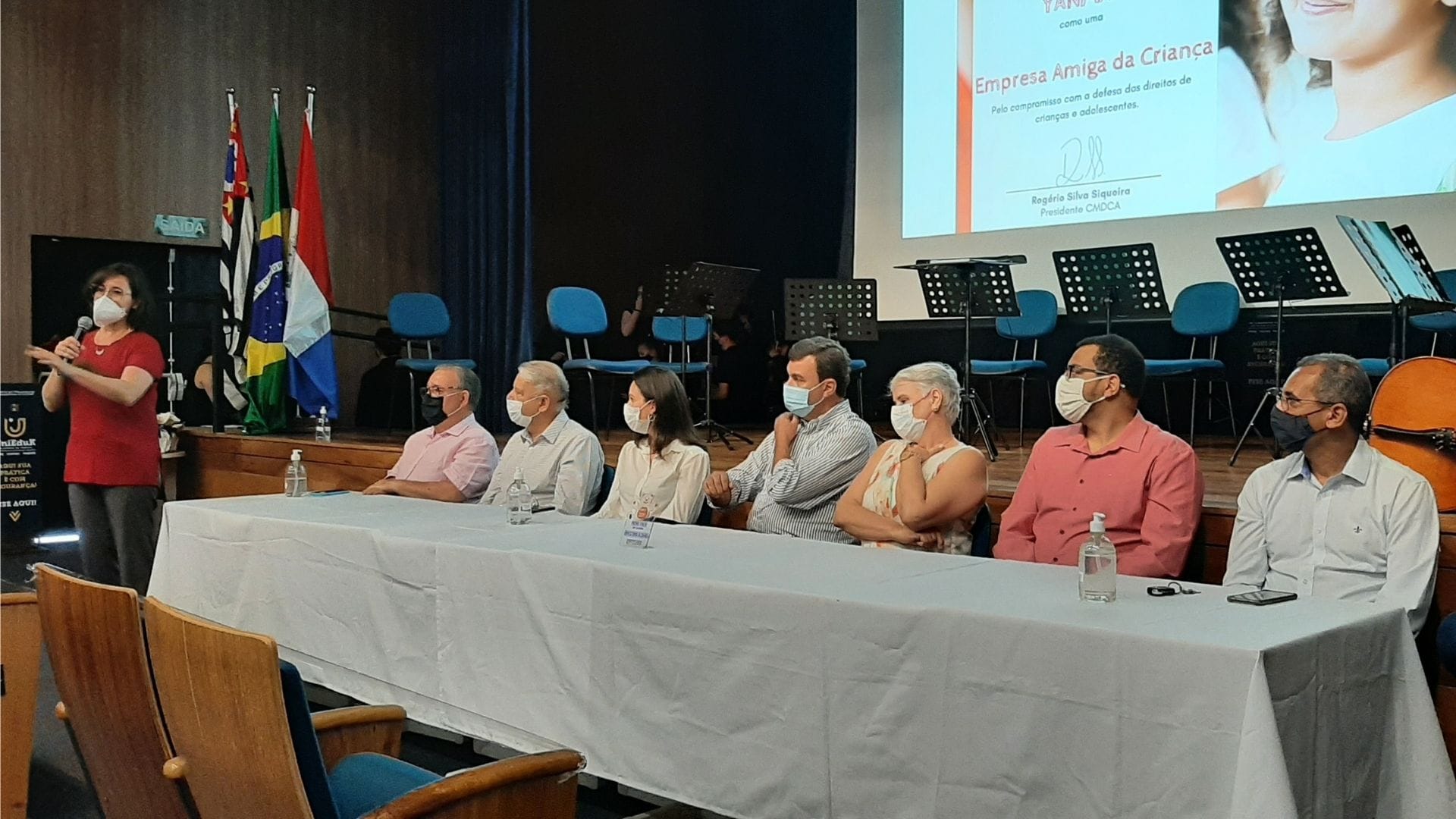 Imagem mostra a banca de autoridades com o Prefeito de Indaiatuba e a coordenadora da UniMAX, e a docente Carla Borges discursando em pé.