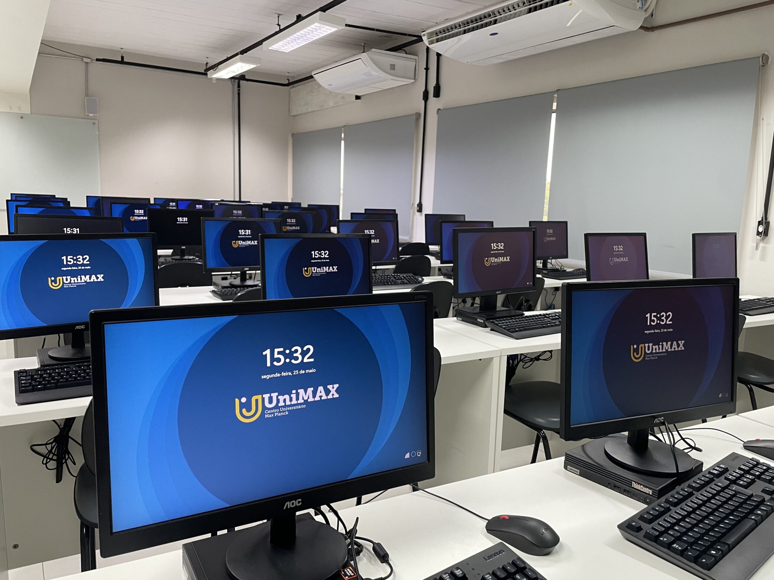 Laboratório de informática da UniMAX com dezenas de computadores organizados em fileiras sobre mesas brancas. Todos os monitores exibem a identidade visual da instituição, em um ambiente amplo, climatizado e preparado para atividades práticas de tecnologia.