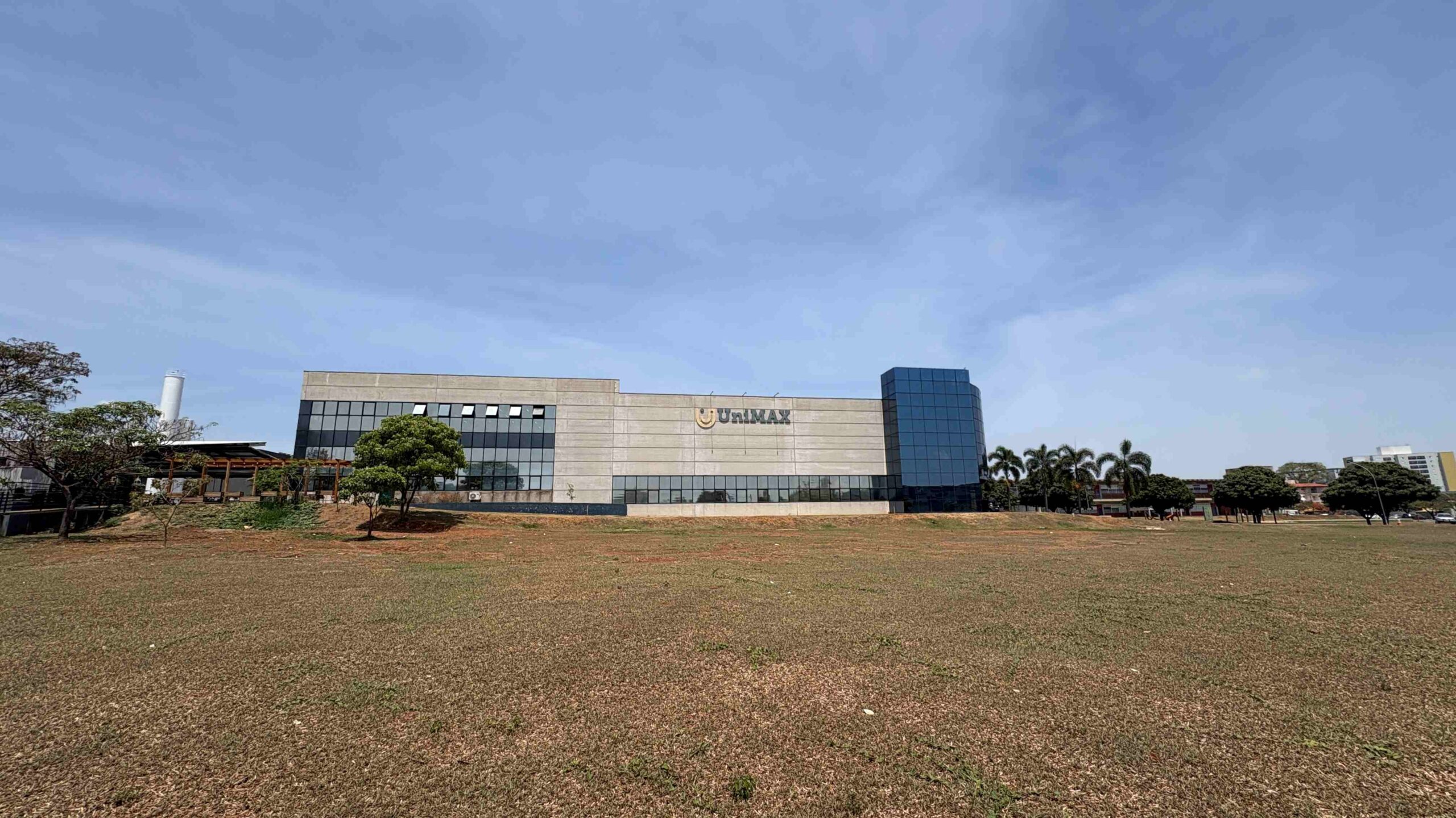 Vista externa de um dos prédios da UniMAX. A construção moderna tem fachada em tons de cinza, grandes janelas de vidro e o logotipo da instituição em destaque. À frente, há um amplo gramado e algumas árvores. O céu azul, com muitas nuvens, ocupa grande parte da imagem, valorizando a paisagem do campus.