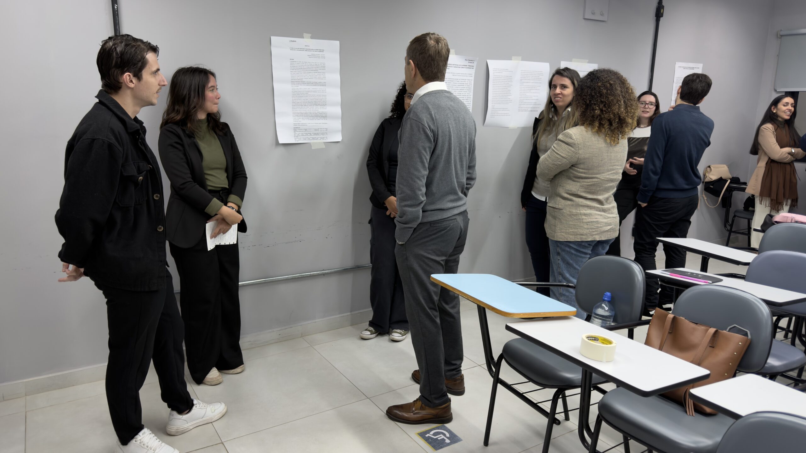 Em uma sala de aula, estudantes de Direito apresentam pesquisas em pôsteres fixados na parede. Professores e colegas conversam com os autores dos trabalhos, observando e debatendo os temas apresentados em um ambiente acadêmico.