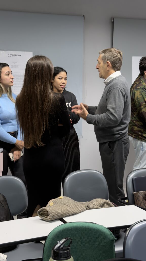 Em uma sala de aula, estudantes de Direito apresentam pesquisas em pôsteres fixados na parede. Professores e colegas conversam com os autores dos trabalhos, observando e debatendo os temas apresentados em um ambiente acadêmico.
