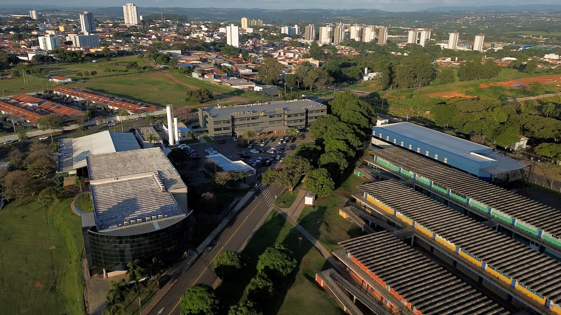 Vista aérea do campus da UniMAX, em Indaiatuba, mostrando os prédios da instituição cercados por áreas verdes, vias internas e estacionamento. Ao fundo, aparecem bairros da cidade e edifícios residenciais sob céu parcialmente aberto.