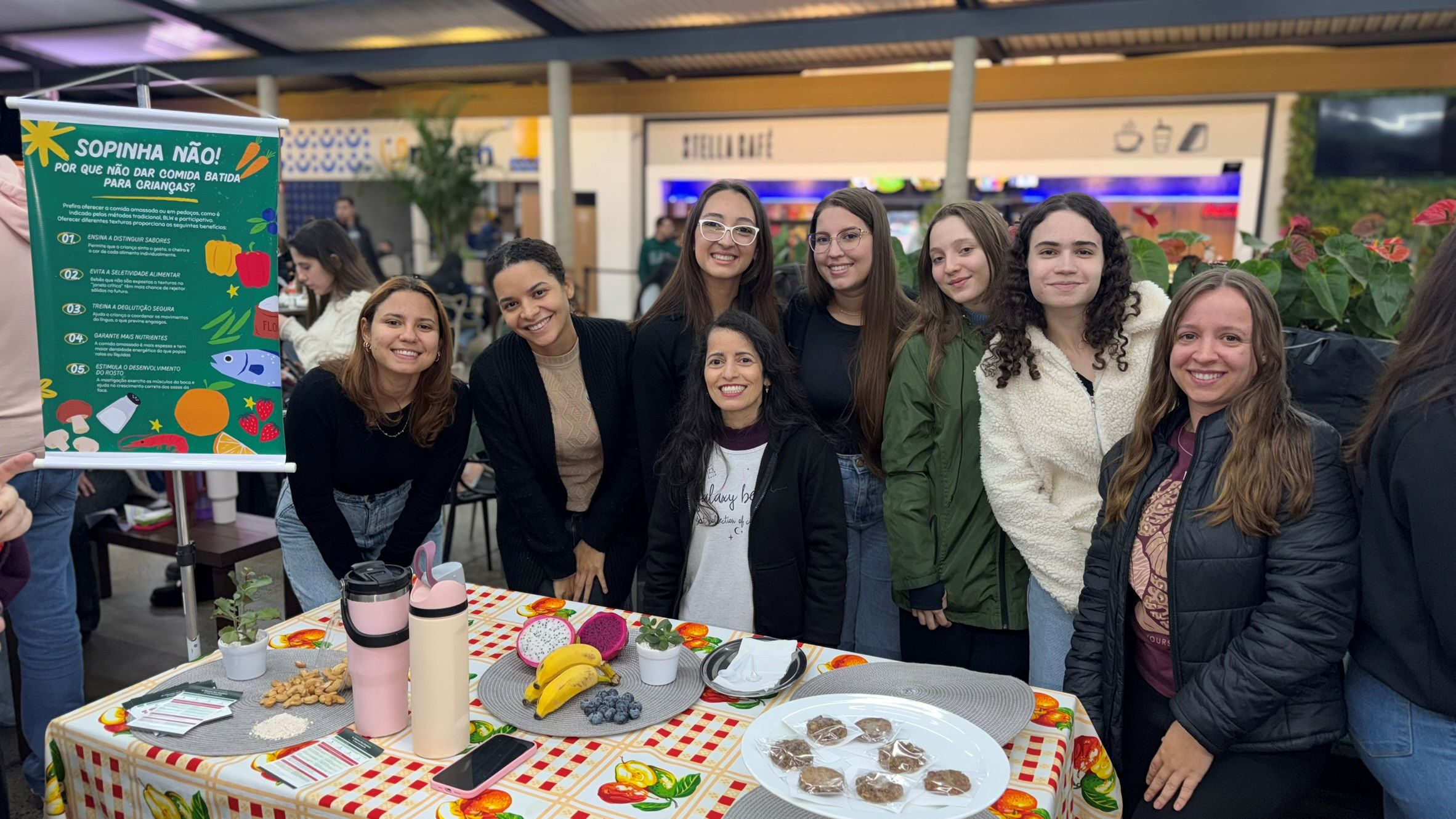 Estudantes do curso de Nutrição posam ao redor de um estande educativo. Sobre a mesa há frutas, alimentos, materiais informativos e exemplos de preparações. Ao lado, um banner destaca orientações sobre alimentação infantil e a importância dos primeiros 1.100 dias de vida.