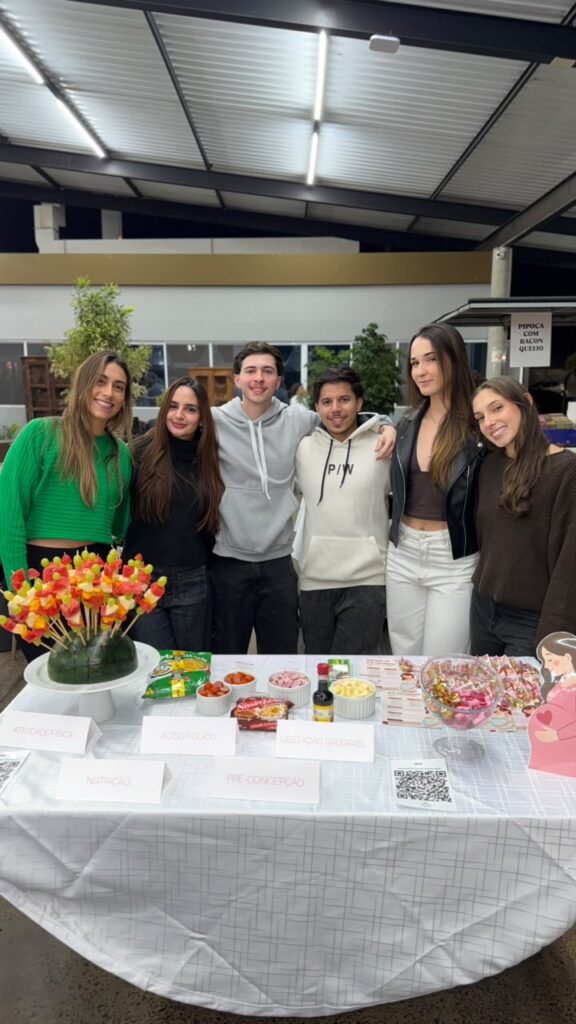 Estudantes do curso de Nutrição posam ao redor de um estande educativo. Sobre a mesa há frutas, alimentos, materiais informativos e exemplos de preparações. Ao lado, um banner destaca orientações sobre alimentação infantil e a importância dos primeiros 1.100 dias de vida.