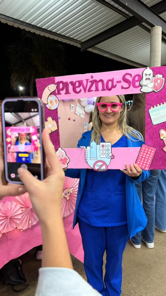 Em um estande decorado com balões nas cores branca e lilás, estudantes de Enfermagem conversam com um participante durante uma ação educativa sobre saúde sexual. Sobre a mesa há materiais informativos e recursos utilizados para orientar sobre prevenção de ISTs e métodos contraceptivos.