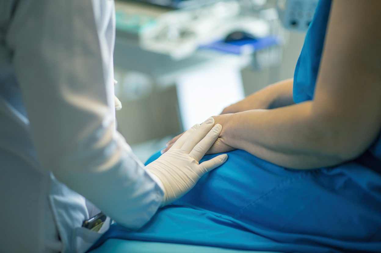 Em um ambiente hospitalar, um profissional de saúde, usando jaleco e luvas, segura delicadamente a mão de um paciente sentado em uma maca. A imagem simboliza acolhimento, cuidado e apoio durante o atendimento em saúde mental.